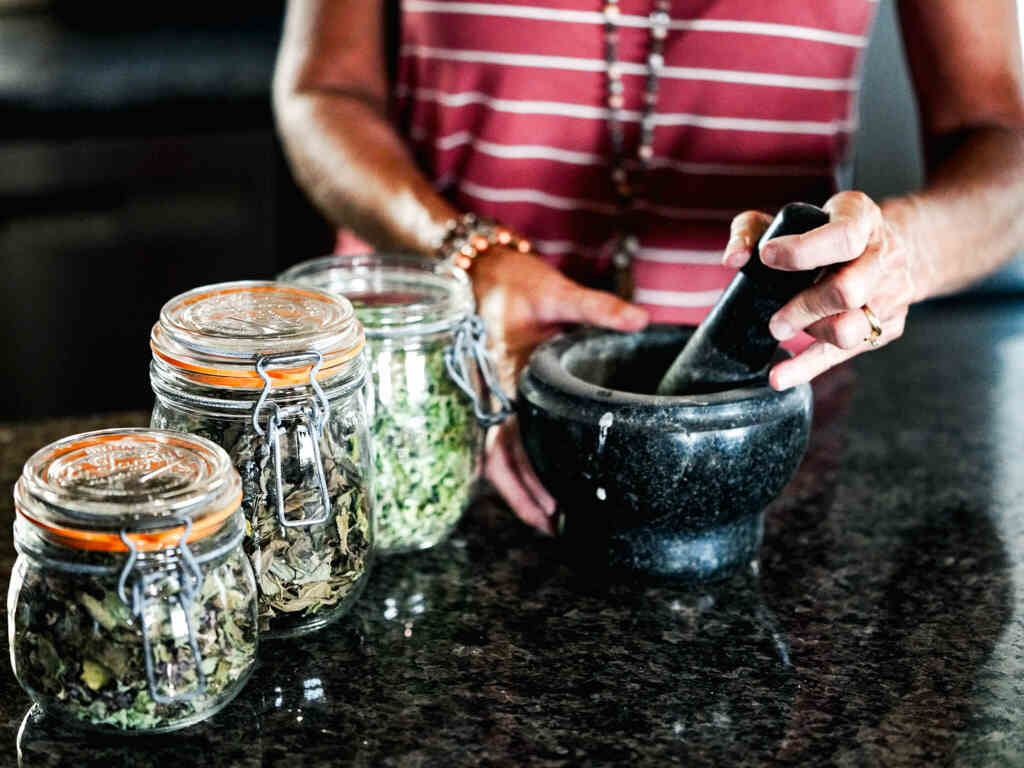 a gal in pink and white striped shirt grinding herbs with black stone mill and glass jars of herbs sitting on black counter.