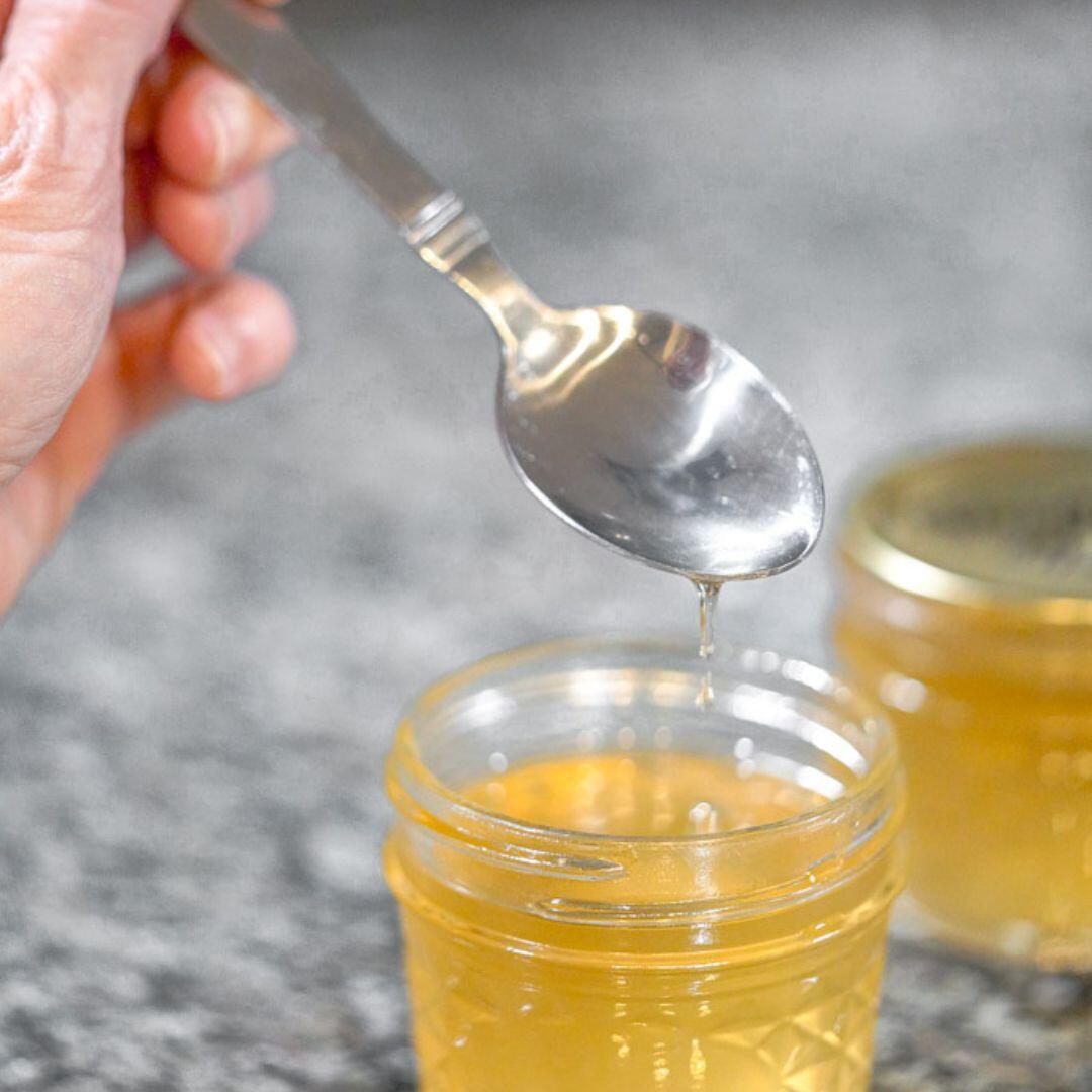 spoon pouring elderflower syrup in small glass jars on black marbled countertop.