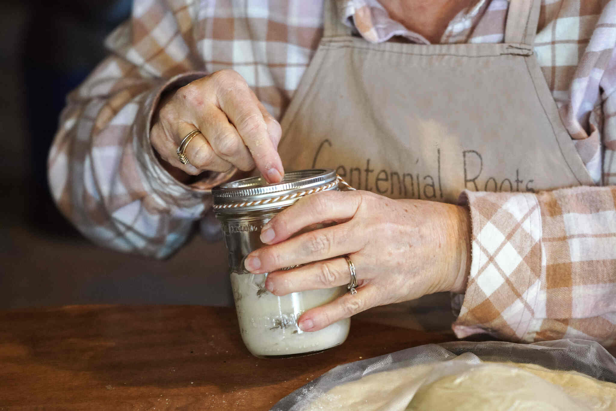 gal in plaid long-sleeve shirt with tan apron stirring sourdough starter