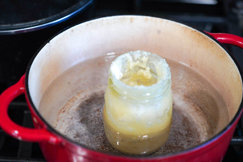 red Dutch oven with water and glass jar of beeswax mixture in it on stove.